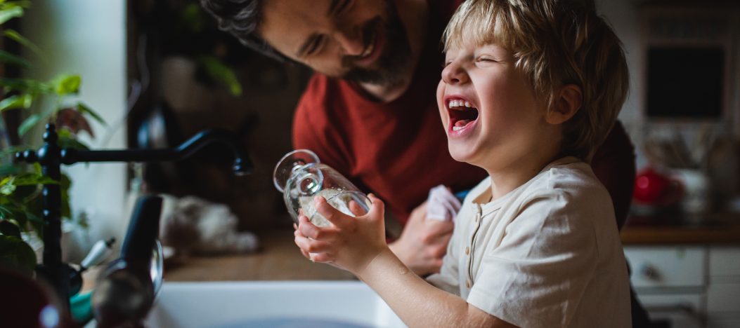 Petit garçon riant avec un père s'amusant en lavant la vaisselle à l'intérieur à la maison, concept de tâches quotidiennes.